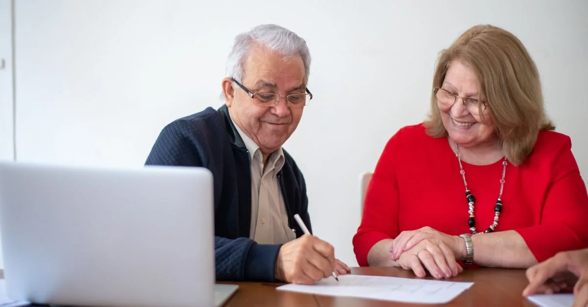 Pareja de ancianos firmando documentos en un escritorio con una laptop. Sonriendo y trabajando.