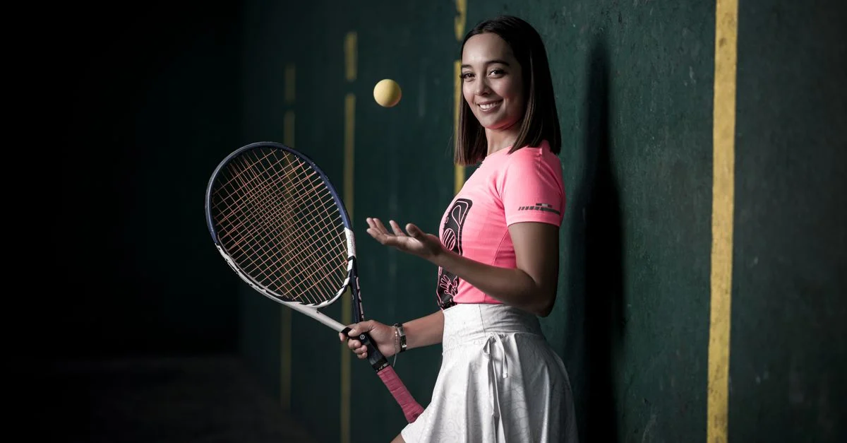 Mujer sonriente posando con racket de tenis y una pelota en interiores, lista para el juego.