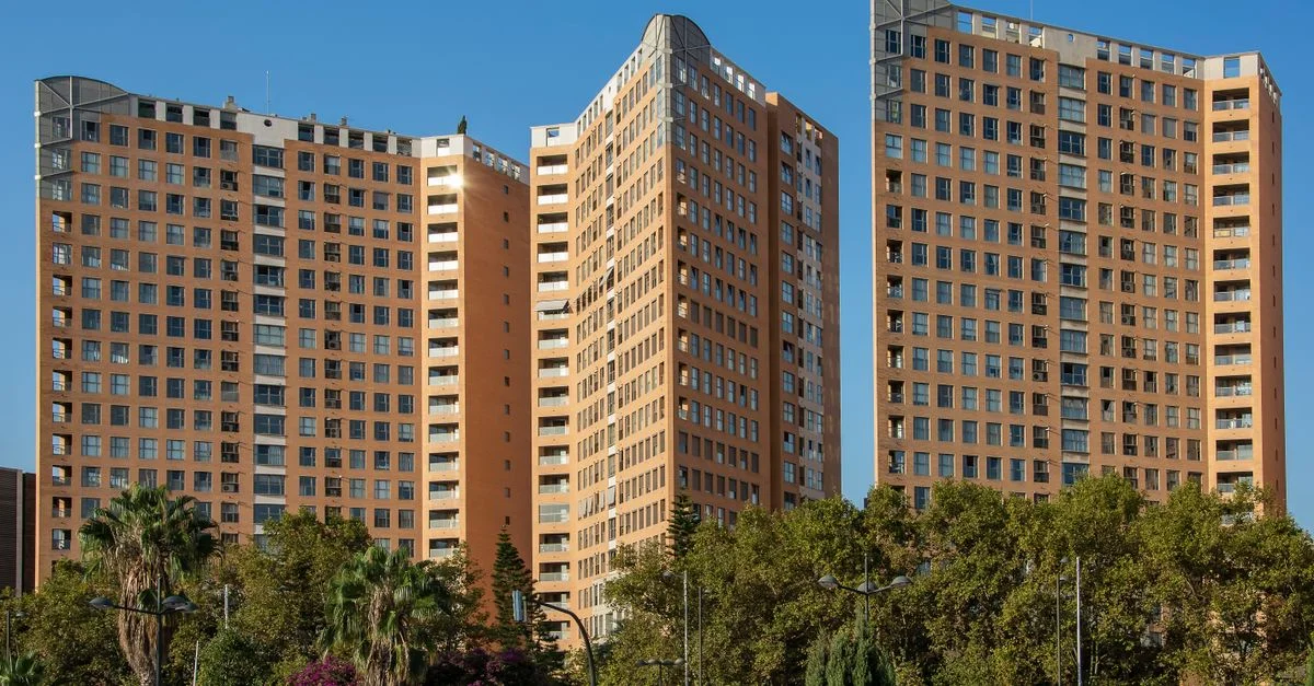 Tres modernos edificios de apartamentos altos se alzan contra un cielo azul claro, rodeados de vegetación exuberante.