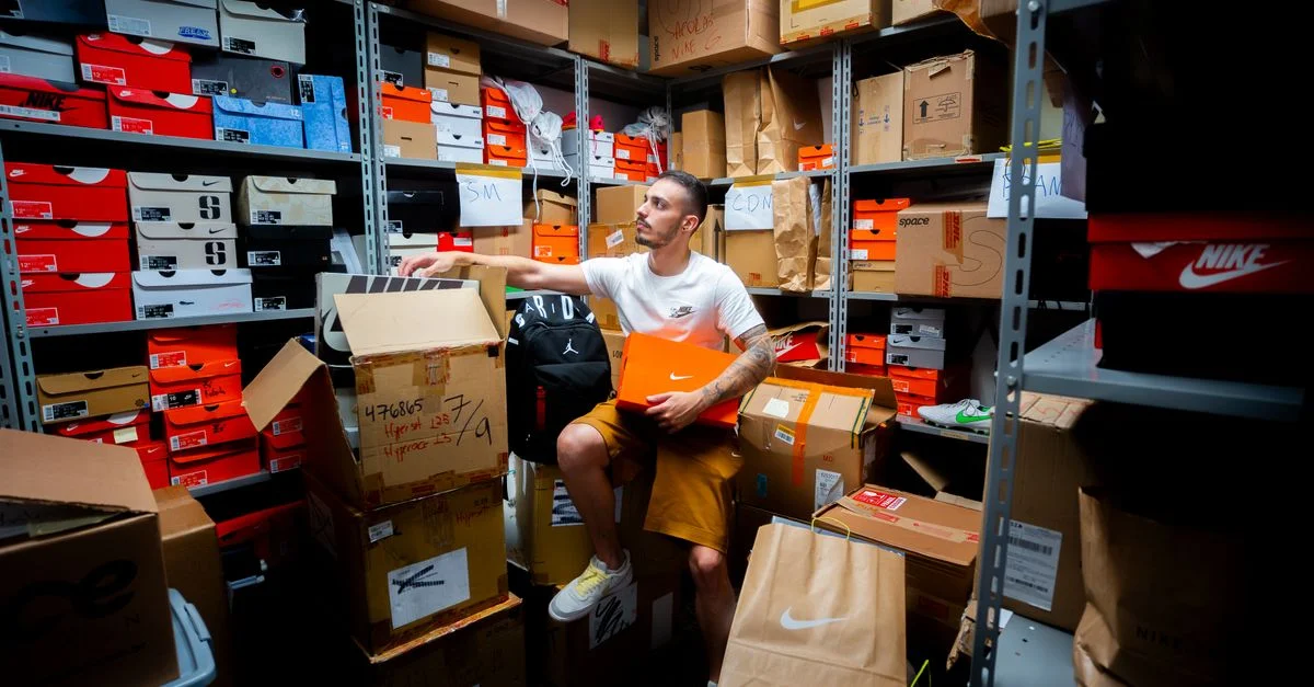 Un hombre organizando varias cajas de zapatos en un almacén desordenado lleno de estanterías y cartones.
